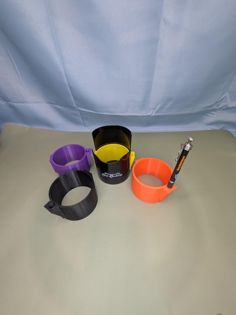 Op de afbeelding: Vier cilindervormige bekerhouders in paars, zwart, geel en oranje. De zwarte houder is voorzien van de tekst Magnetic Dip Caddy. Een oranje houder heeft een zwarte pen met een zilveren clip in een kleine zijbevestiging aan de rand gestoken.