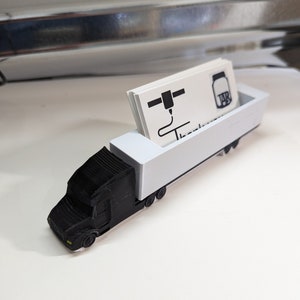 May include: A black and white 3D printed truck business card holder. The truck is a semi-trailer truck with a white trailer. The business card holder is designed to hold business cards in the trailer. The business card in the holder has a 3D printer icon and the text "TAB"