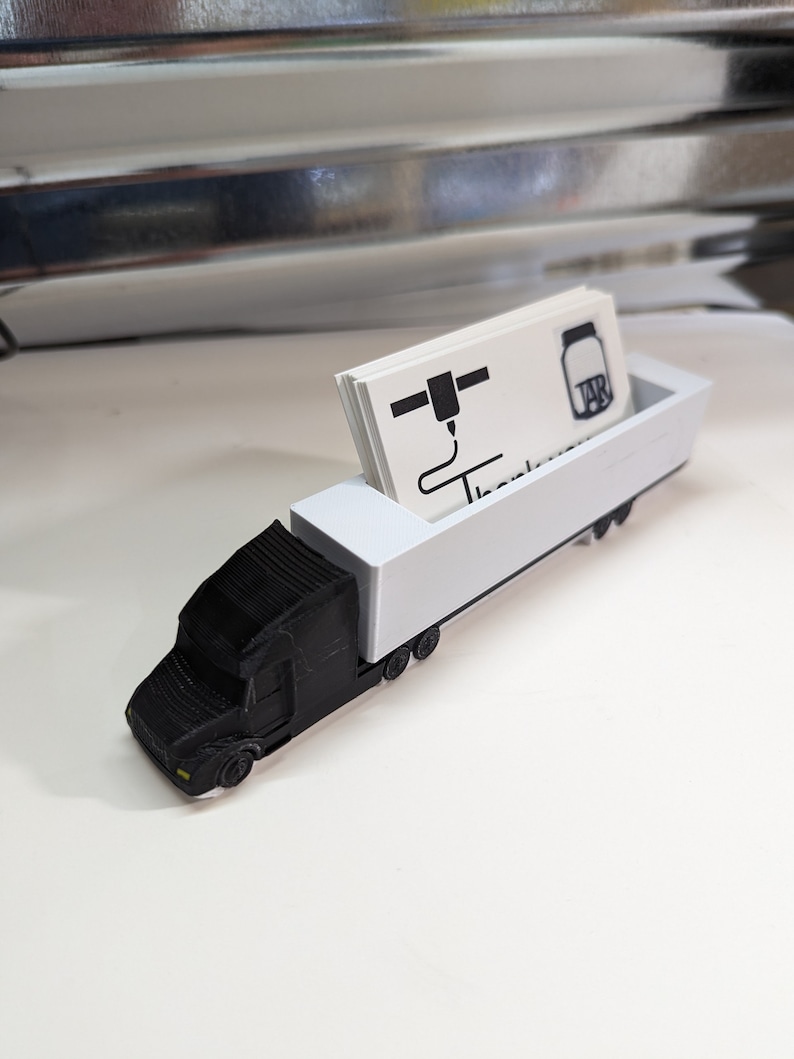 May include: A black and white 3D printed truck business card holder. The truck is a semi-trailer truck with a white trailer. The business card holder is designed to hold business cards in the trailer. The business card in the holder has a 3D printer icon and the text "TAB"