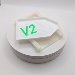 Puede incluir: Bandeja de pintura de diamantes blanca con una base giratoria blanca. La bandeja tiene una superficie texturizada y el texto "V2" est&aacute; impreso en la bandeja.