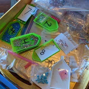 Puede incluir: Primer plano de una bandeja llena de diversos materiales para la pintura de diamantes. La bandeja contiene peque&ntilde;os recipientes de pl&aacute;stico llenos de diamantes de imitaci&oacute;n de colores, etiquetados con n&uacute;meros. Tambi&eacute;n hay varias bolsas de pl&aacute;stico transparentes que contienen diamantes de imitaci&oacute;n, una herramienta de forma de diamante rosa y una herramienta verde con mango.