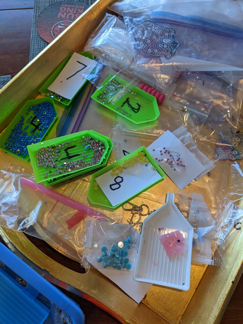 Puede incluir: Primer plano de una bandeja llena de diversos materiales para la pintura de diamantes. La bandeja contiene peque&ntilde;os recipientes de pl&aacute;stico llenos de diamantes de imitaci&oacute;n de colores, etiquetados con n&uacute;meros. Tambi&eacute;n hay varias bolsas de pl&aacute;stico transparentes que contienen diamantes de imitaci&oacute;n, una herramienta de forma de diamante rosa y una herramienta verde con mango.