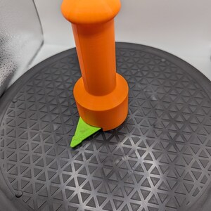 May include: An orange and green 3D printed tool with a round top and a pointed base. The tool is sitting on a black circular surface with a triangular pattern.