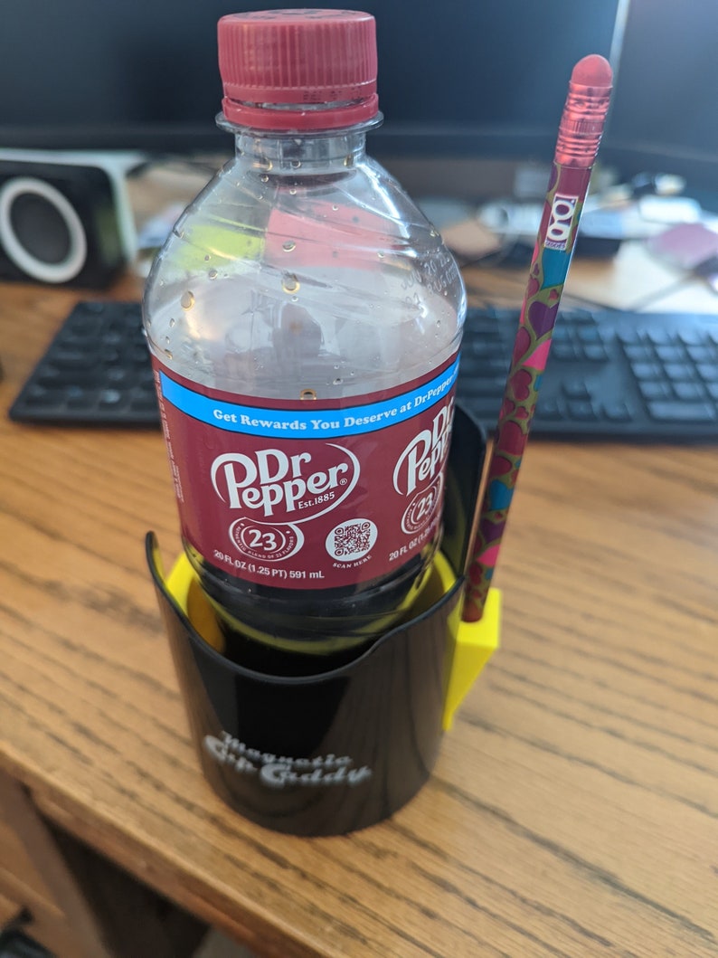 Op de afbeelding: Een zwarte plastic bekerhouder met een gele zijbevestiging met daarin een Dr Pepper fles van 591 ml en een potlood met patroon. De bekerhouder is voorzien van de tekst Magnetic Sip Caddy en het flesetiket toont het Dr Pepper merk.