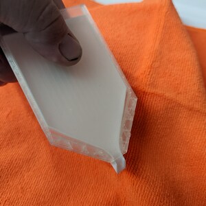 Puede incluir: Un objeto blanco de forma hexagonal impreso en 3D con una superficie texturizada. El objeto est&aacute; siendo sostenido por una mano y descansa sobre un pa&ntilde;o de microfibra de color naranja brillante.