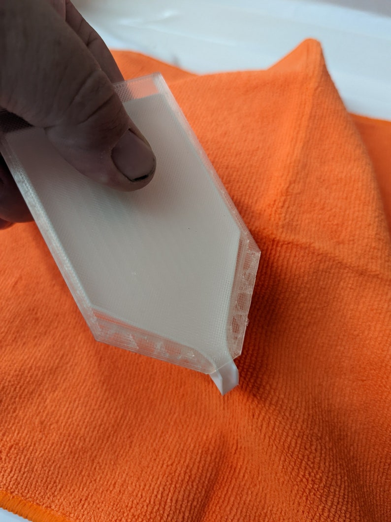 Puede incluir: Un objeto blanco de forma hexagonal impreso en 3D con una superficie texturizada. El objeto est&aacute; siendo sostenido por una mano y descansa sobre un pa&ntilde;o de microfibra de color naranja brillante.