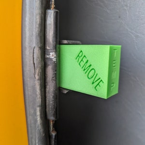 May include: A green plastic sign with the word "REMOVE" printed on it. The sign is attached to a black metal hinge.