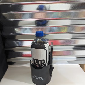 Op de afbeelding: Een zwarte kunststof Magnetic Cup Caddy bekerhouder met een doorzichtige plastic fles met een blauwe dop en een wit etiket met de tekst TAR. Een zwarte pen met zilverkleurige accenten is bevestigd aan de zijkant van de bekerhouder voor dranken.