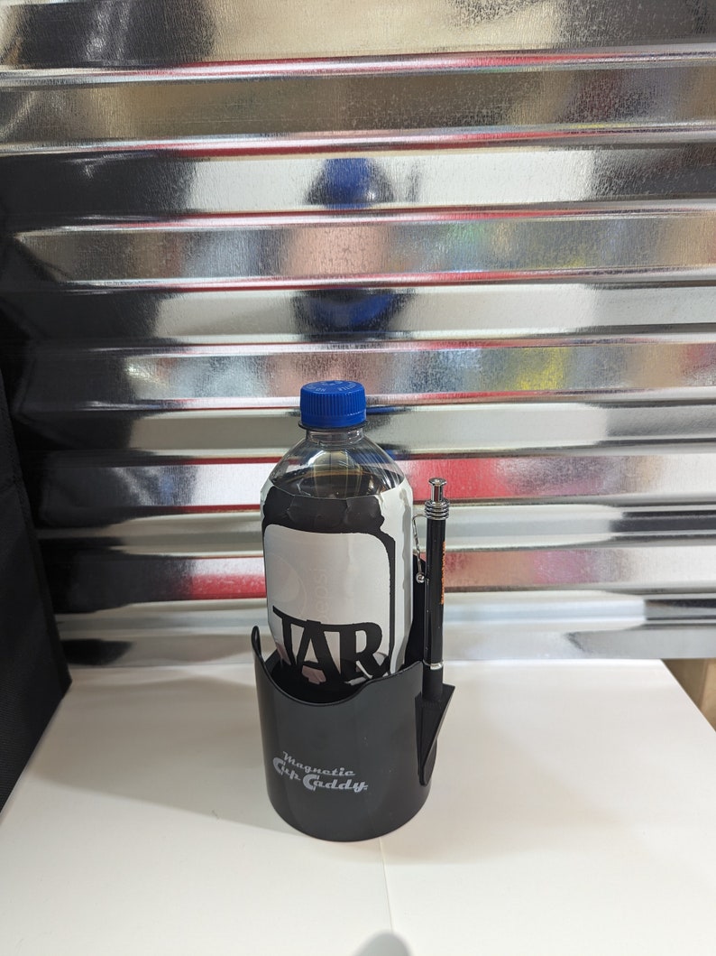 Op de afbeelding: Een zwarte kunststof Magnetic Cup Caddy bekerhouder met een doorzichtige plastic fles met een blauwe dop en een wit etiket met de tekst TAR. Een zwarte pen met zilverkleurige accenten is bevestigd aan de zijkant van de bekerhouder voor dranken.