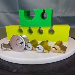 May include: A collection of locksmith tools and components, including a silver lock, several brass cylinders, and keys. A bright green and yellow block structure is in the background, providing a visual contrast. The items are arranged on a white circular surface.