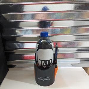 Op de afbeelding: Een zwarte Magnetic Cup Caddy bekerhouder met oranje accenten, met daarin een doorzichtige plastic fles met een blauwe dop en een zwart etiket met de tekst TAB. Een zwart-zilveren vulpotlood is in de zijkant van de bekerhouder gestoken.