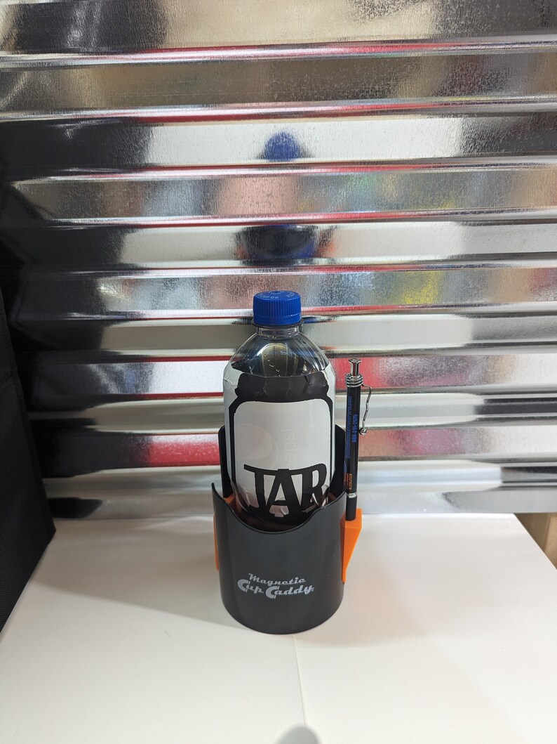 Op de afbeelding: Een zwarte Magnetic Cup Caddy bekerhouder met oranje accenten, met daarin een doorzichtige plastic fles met een blauwe dop en een zwart etiket met de tekst TAB. Een zwart-zilveren vulpotlood is in de zijkant van de bekerhouder gestoken.