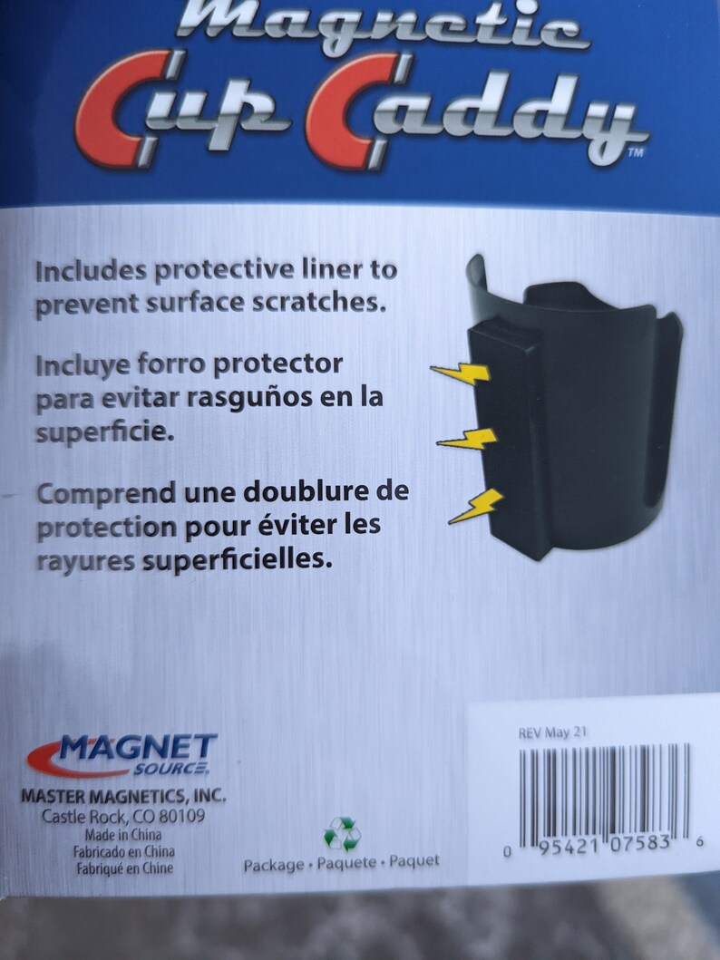 Op de afbeelding: Een zwarte magnetische bekerhouder met een beschermende voering om krassen op oppervlakken te voorkomen. De verpakking toont de productnaam, drie gele bliksemschichtpictogrammen die naar de magneet wijzen en een barcode met nummer 0 95421 07583 6.