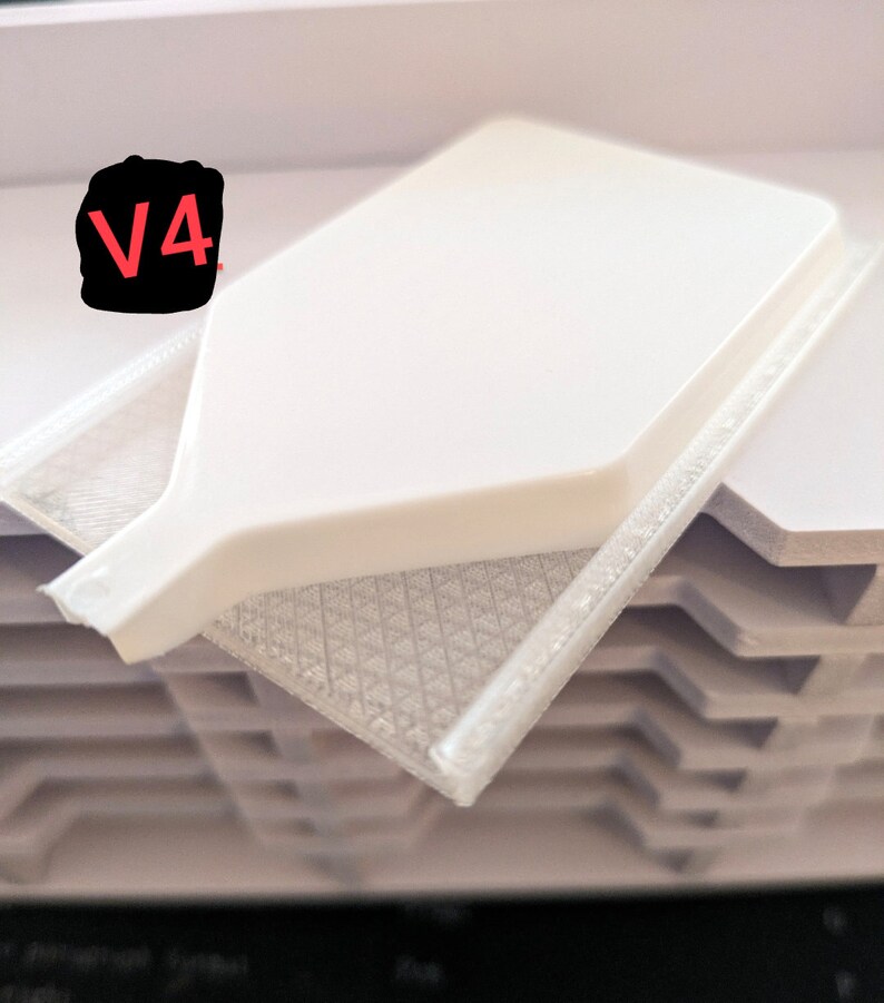 Puede incluir: Un objeto blanco de pl&aacute;stico impreso en 3D con una etiqueta V4. El objeto tiene forma de embudo con un pico largo. El objeto est&aacute; colocado sobre una pila de l&aacute;minas de pl&aacute;stico blancas.