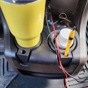May include: A black car cup holder with a yellow travel mug, a white bottle, and a yellow pen. The cup holder is attached to the car's center console.