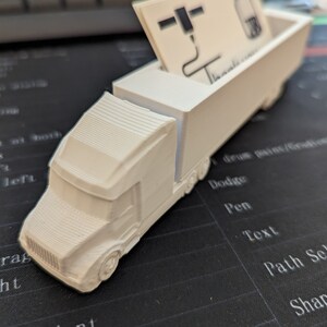 May include: White plastic truck shaped business card holder. The truck has a flatbed with a slot for business cards. The business card in the slot is white with black text and a logo.