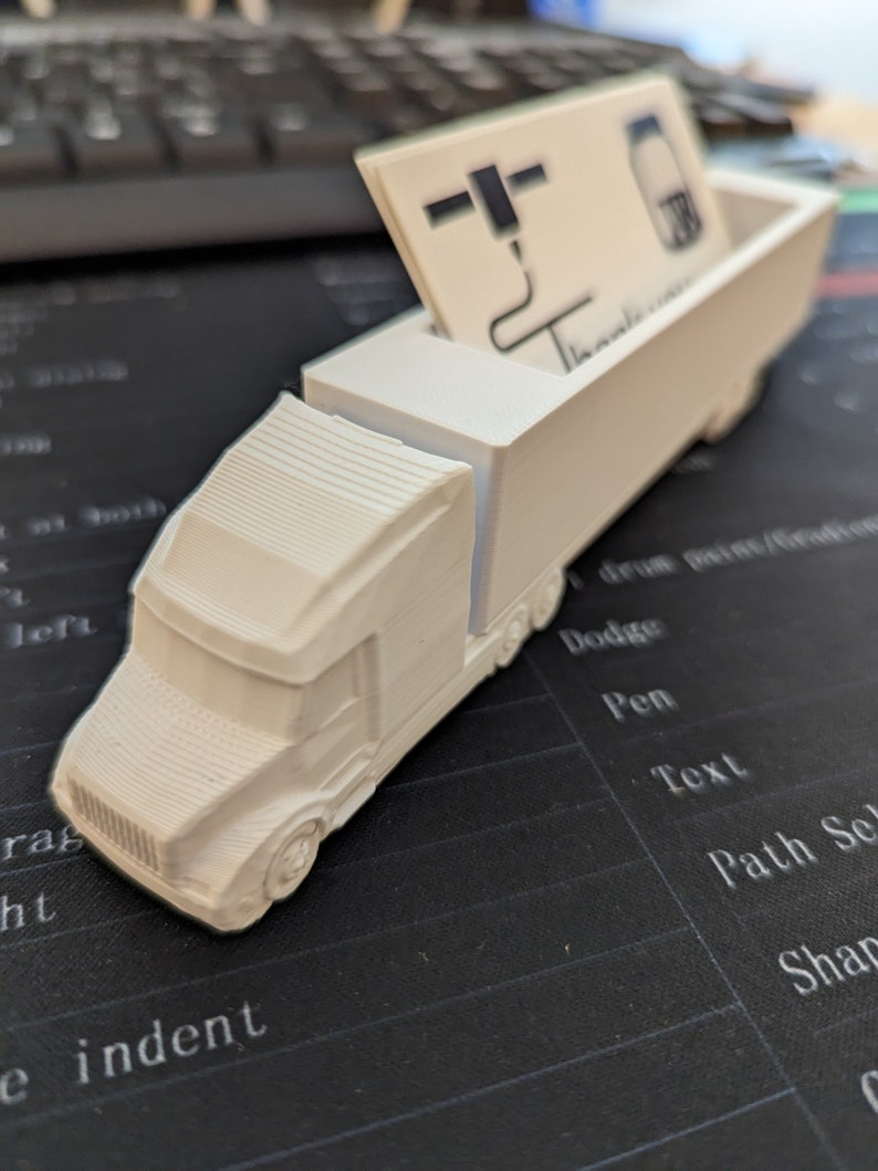 May include: White plastic truck shaped business card holder. The truck has a flatbed with a slot for business cards. The business card in the slot is white with black text and a logo.