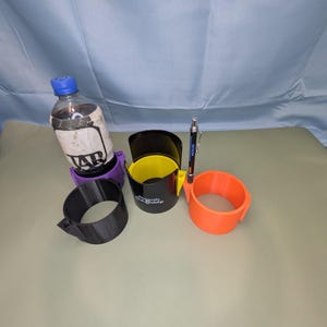 Op de afbeelding: Een verzameling van vier cilindrische bekerhouders in paars, zwart, geel en oranje. E&eacute;n paarse houder bevat een plastic fles met een etiket waarop JAR staat. De zwarte houder bevat de tekst Magnetic Cup Buddy. Een pen is in de oranje houder gestoken.