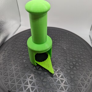 May include: A bright green 3D printed bottle opener with a pointed tip and a cylindrical handle. The opener is sitting on a black surface with a triangular pattern.