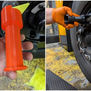May include: A bright orange cylindrical tool with a yellow triangular tip designed for vehicle maintenance. The tool features a flared base and a hollow top section, shown in use to align or secure lug nuts on a large commercial vehicle wheel.
