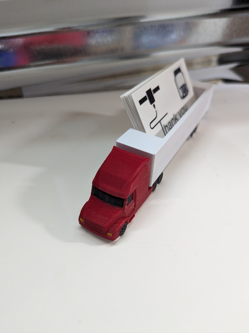 May include: A red toy semi-truck with a white trailer. The trailer has a slot for holding business cards. The business cards have a logo and the text "Thank you".