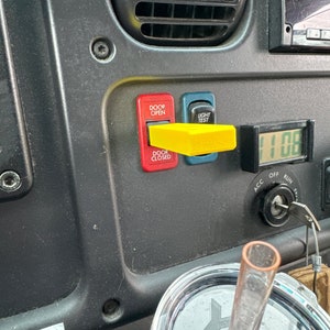 May include: Close-up of a truck's dashboard with red and yellow buttons labeled "Door Open" and "Light Test". A yellow plastic lever is in the "Light Test" position. A digital clock displays "11:08".