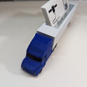 May include: A blue toy semi-truck with a white trailer. The trailer has a slot for holding business cards. The business cards have a black and white logo and the text "Thank You".