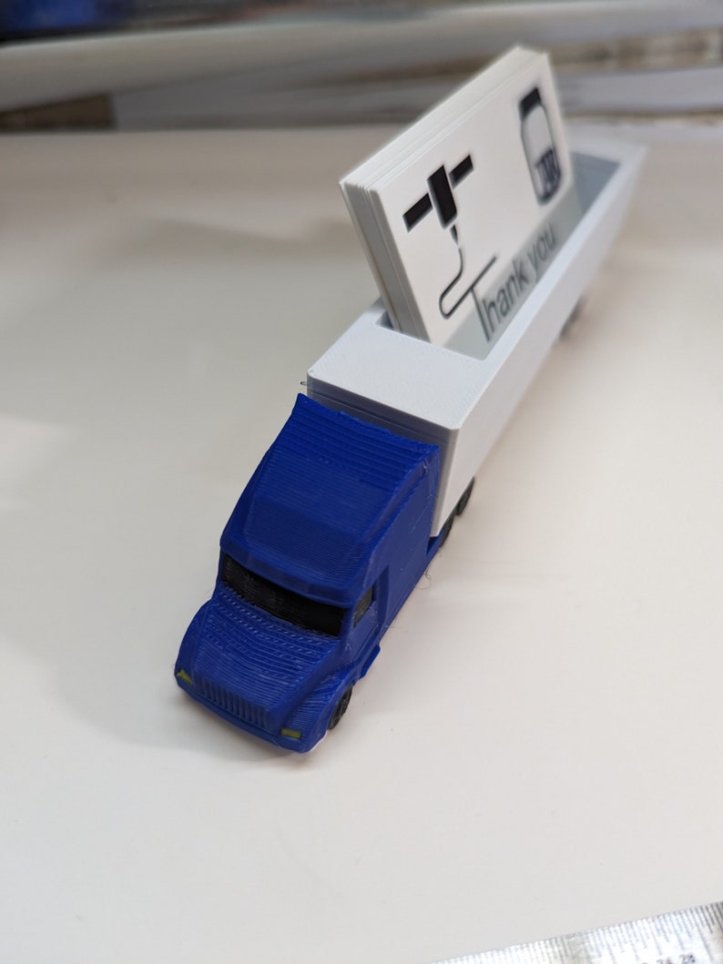 May include: A blue toy semi-truck with a white trailer. The trailer has a slot for holding business cards. The business cards have a black and white logo and the text "Thank You".