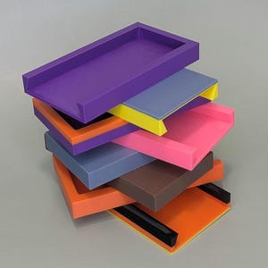Puede incluir: Una pila de seis bandejas de pl&aacute;stico de colores con bordes de diferentes colores. Las bandejas est&aacute;n apiladas en forma escalonada, con la bandeja superior de color morado y la bandeja inferior de color naranja.