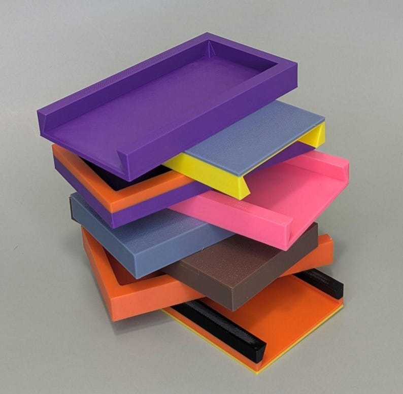 Puede incluir: Una pila de seis bandejas de pl&aacute;stico de colores con bordes de diferentes colores. Las bandejas est&aacute;n apiladas en forma escalonada, con la bandeja superior de color morado y la bandeja inferior de color naranja.