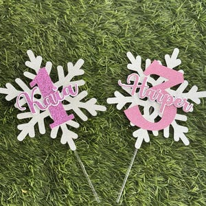 Peut inclure: Deux d&eacute;corations de g&acirc;teau en forme de flocon de neige blanc avec des paillettes roses. Un topper porte le num&eacute;ro "1" et le nom "Kaiya". L'autre topper porte le num&eacute;ro "2" et le nom "Harper".