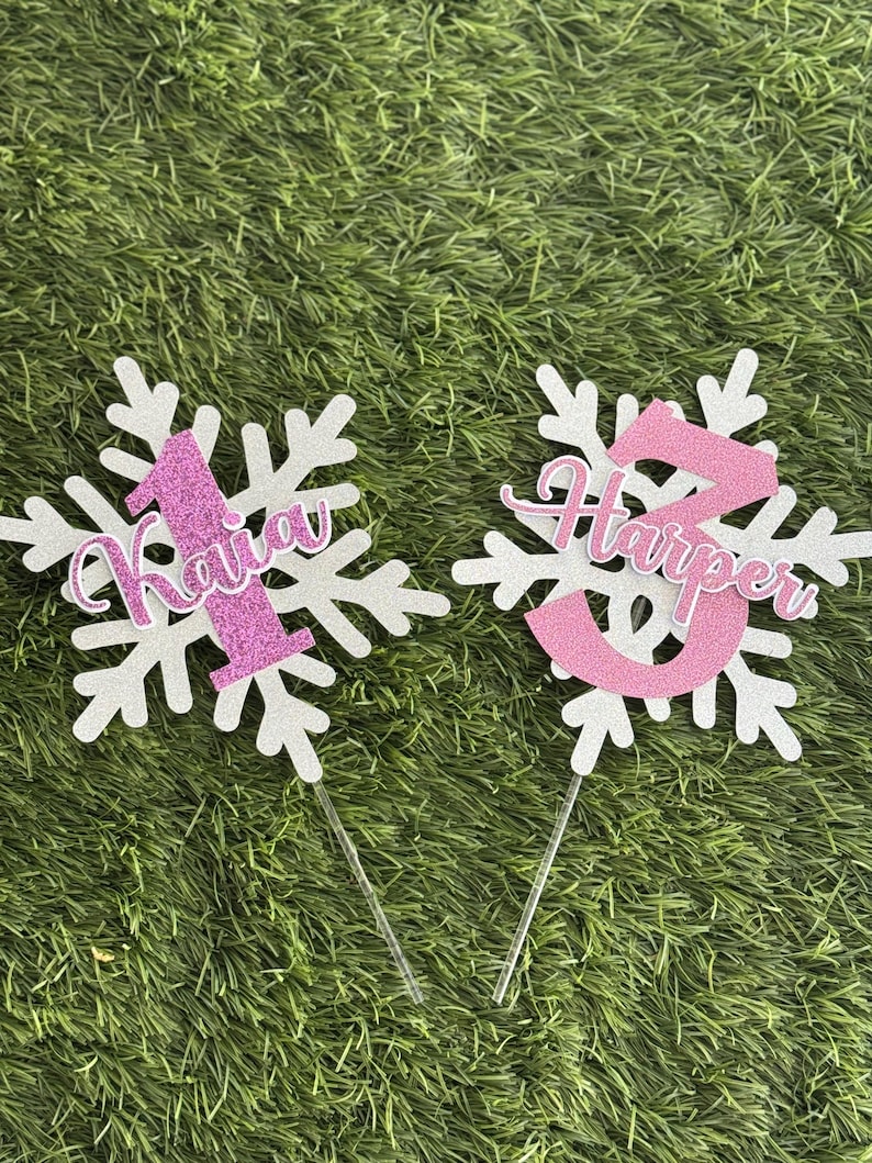 Peut inclure: Deux d&eacute;corations de g&acirc;teau en forme de flocon de neige blanc avec des paillettes roses. Un topper porte le num&eacute;ro "1" et le nom "Kaiya". L'autre topper porte le num&eacute;ro "2" et le nom "Harper".