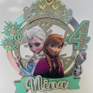 Peut inclure: Un topper de g&acirc;teau repr&eacute;sentant deux princesses Disney, Elsa et Anna, avec une couronne scintillante, des flocons de neige et le chiffre 4. Le topper comprend &eacute;galement le nom "Nina" en lettres vertes scintillantes.