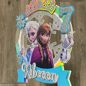 Peut inclure: Un topper de g&acirc;teau en paillettes argent&eacute;es avec une couronne, un flocon de neige et le chiffre 7. Le topper pr&eacute;sente &eacute;galement des images d'Elsa et d'Anna de la Reine des Neiges, ainsi que le nom "Rebecca".