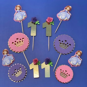 May include: Eleven cupcake toppers featuring a princess theme. The toppers include two gold numbers "1", two gold numbers "1" with pink flowers, two purple crowns, two princesses in purple dresses, and one princess in a blue dress.