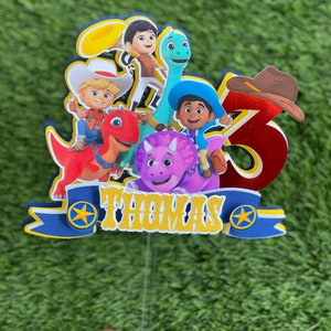 May include: A multi-layered cake topper featuring cartoon characters, a red dinosaur, a purple triceratops, and a blue long-necked dinosaur. It includes a large red number 3 with a brown cowboy hat and a blue banner with the name THOMAS in gold.