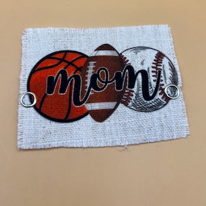 May include: A white fabric patch with a basketball, football, and baseball design. The word "MOM" is written in black cursive across the balls.