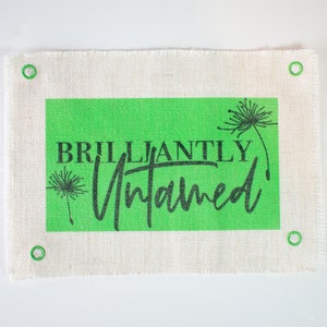 May include: A white canvas fabric with green and black text that reads "BRILLIANTLY Untamed" with a dandelion graphic. The fabric has four green grommets around the edges.