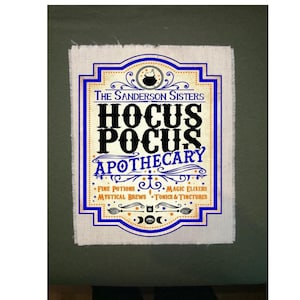May include: A rectangular sign with a blue and gold border, featuring the text "The Sanderson Sisters Hocus Pocus Apothecary." The design includes illustrations of a cauldron, stars, and other mystical elements. The sign is on a textured, off-white background.