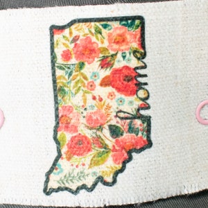 May include: A white fabric patch with a black outline of the state of Indiana. The patch is covered in a floral pattern with pink, red, and green flowers.