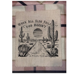 May include: A black and white desert landscape illustration on a beige fabric. The image features a road leading to a setting sun, surrounded by cacti. Text reads "WHEN ALL ELSE FAILS GOD DOESN'T" and "PSALMS 73:26".