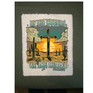 May include: A fabric patch featuring a desert landscape with cacti, a cross, and a setting sun. The text "BY HIS WOUNDS, WE ARE HEALED" is displayed above and below the cross, with the Bible verse 1 Peter 2:24 at the bottom.