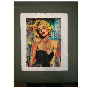 May include: A framed art print featuring a portrait of a woman with blonde hair and red lipstick. The artwork has a distressed, graffiti-style background with vibrant colors and text. The woman is wearing a black dress.