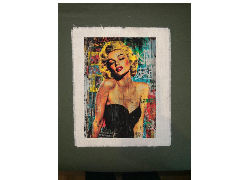 May include: A framed art print featuring a portrait of a woman with blonde hair and red lipstick. The artwork has a distressed, graffiti-style background with vibrant colors and text. The woman is wearing a black dress.