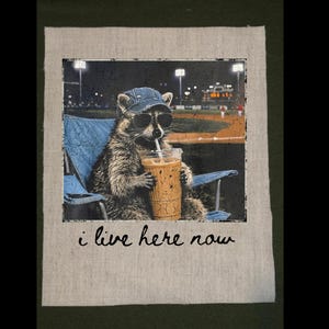 May include: A raccoon wearing sunglasses and a denim cap, sitting in a blue folding chair, holding a drink with a straw. The background shows a baseball field at night. The text "i live here now" is below the image.