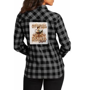 May include: Black and gray plaid flannel shirt with a "Cowgirl Rodeo Nights" graphic on the back. The graphic features a cowgirl on a horse with desert scenery. The shirt has long sleeves and a classic button-down design.