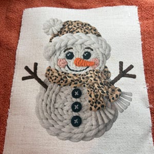 May include: A fabric snowman design with a leopard print hat and scarf. The snowman has a carrot nose, black button eyes, and a smiling mouth. The snowman's body is made of gray yarn and has three black buttons.