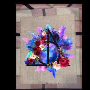 May include: A decorative design featuring a black triangle and circle symbol, adorned with red, white, and pink roses, set against a backdrop of blue and purple watercolor-style splashes. The design is on a beige fabric.