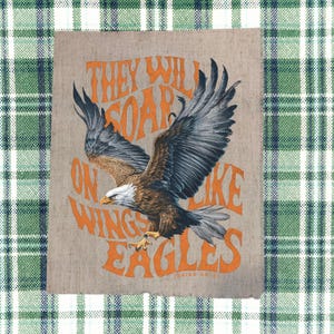 May include: A print featuring a detailed illustration of a bald eagle with outstretched wings. The text "THEY WILL SOAR ON WINGS LIKE EAGLES" is in orange. The background is a neutral tan color.