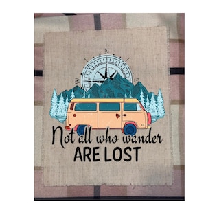 May include: A tan and orange camper van illustration with a compass and mountain scene. The text "Not all who wander are lost" is below the van. The image is on a tan fabric with a plaid border.